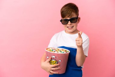 Küçük kızıl saçlı çocuk pembe arka planda gözlüklü ve elinde büyük bir kova patlamış mısırla ön tarafı işaret ediyor.