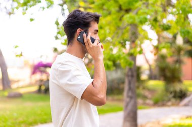 Parkta genç beyaz bir adam cep telefonuyla biriyle sohbet ediyor.