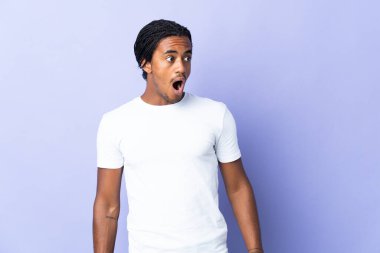 Örgülü genç Afrikalı Amerikalı adam mor arka planda izole edilmiş. Yan tarafa bakarken sürpriz jest yapıyor.