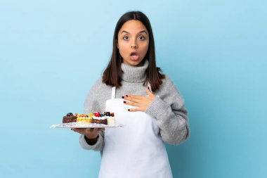 Pasta şefi elinde büyük bir pasta tutuyor mavi arka planda şaşırmış ve şaşırmış..