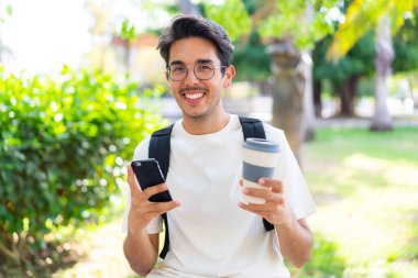 Dışarıda cep telefonu kullanan ve elinde kahve tutan genç bir öğrenci.