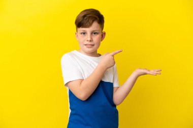 Sarı arka planda izole edilmiş küçük kızıl saçlı çocuk bir ilan vermek için avucunun içinde hayali bir şey tutuyor.