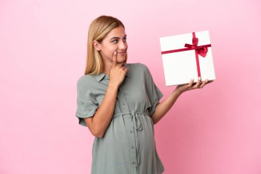 Genç Uruguaylı kadın mavi arka planda izole edilmiş hamile ve elinde şüpheleri varken bir hediye tutuyor.