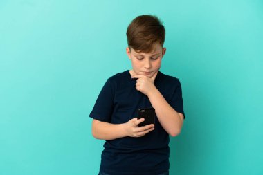 Küçük kızıl saçlı çocuk mavi arka planda izole edilmiş düşünüyor ve bir mesaj gönderiyor.