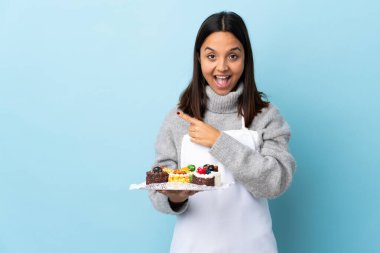 Pasta şefi büyük bir pasta tutuyor. Mavi arka plan üzerinde. Şaşırmış ve işaret ediyor..