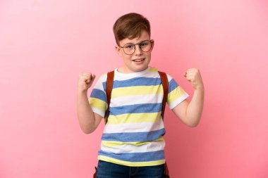 Küçük kızıl saçlı beyaz çocuk pembe arka planda izole edilmiş ve galibiyeti kutluyor.