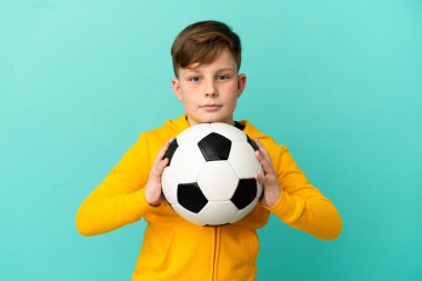 Küçük kızıl saçlı çocuk mavi arka planda futbol topuyla izole edilmiş.