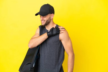 Spor çantalı genç sarışın adam sarı arka planda izole edilmiş, omuz ağrısından muzdarip, çabaladığı için.