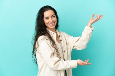 Mavi arka planda izole edilmiş genç beyaz kadın, davet edilmek için ellerini yana uzatıyor.