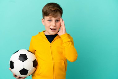 Mavi arka planda izole bir şekilde futbol oynayan kızıl saçlı çocuk ağzı açık bağırıyor.