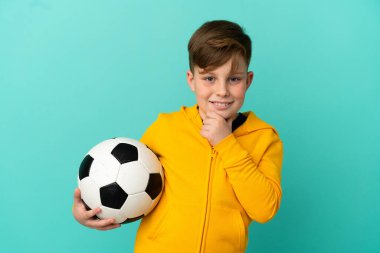 Mavi arka planda izole bir şekilde futbol oynayan kızıl saçlı çocuk yana bakıyor ve gülümsüyor.