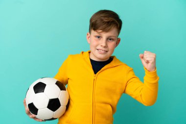 Mavi arka planda izole edilmiş küçük kızıl saçlı çocuk futbol topuyla zaferi kutluyor.