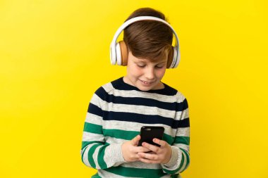 Küçük kızıl saçlı çocuk sarı arka planda izole edilmiş müzik dinliyor ve cep telefonu arıyor.
