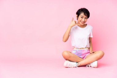 Kısa saçlı genç Vietnamlı kadın izole edilmiş pembe arka planda oturuyor ve telefon jesti yapıyor. Bana geri dönüş işareti ver.