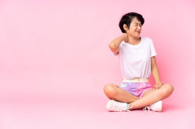 Kısa saçlı, genç Vietnamlı kadın, izole pembe arka planda, boyun ağrısı ile yerde oturuyor.