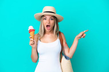 Elinde dondurma ve plaj çantası tutan genç Romen kadın mavi arka planda izole edilmiş şaşırmış ve parmağıyla kenarı işaret ediyor