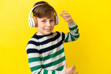 Küçük kızıl saçlı çocuk sarı arka planda izole edilmiş müzik dinliyor ve gitar hareketi yapıyor.