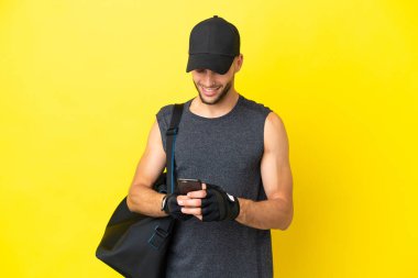 Sarı arka planda izole edilmiş spor çantalı genç sarışın adam cep telefonuyla bir mesaj gönderiyor.
