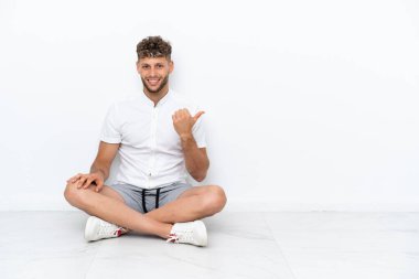 Genç sarışın adam, beyaz arka planda izole bir şekilde yerde oturuyor. Yan tarafı göstererek bir ürün sunuyor.