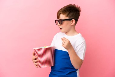 Küçük kızıl saçlı çocuk pembe arka planda 3 boyutlu gözlüklü ve elinde patlamış mısır dolu bir kova tutuyor.