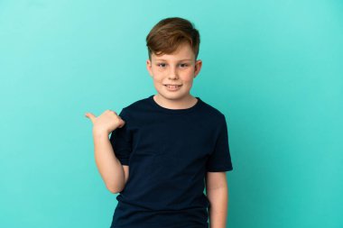 Mavi arka planda izole edilmiş küçük kızıl çocuk bir ürünü sunmak için kenarı işaret ediyor.