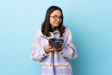 Genç esmer, melez bir kadın elinde bir kase erişteyle izole edilmiş mavi arka planda bir şeyler çeviriyor..