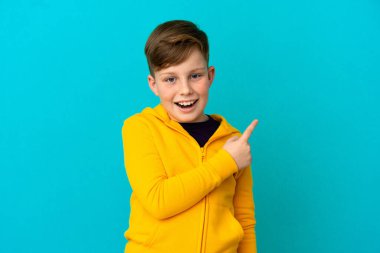 Mavi arka planda izole edilmiş küçük kızıl çocuk şaşırmış ve işaret ediyor.