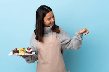 Pasta şefi, izole edilmiş mavi arka planda büyük bir pasta tutuyor ve baş parmak işareti yapıyor..