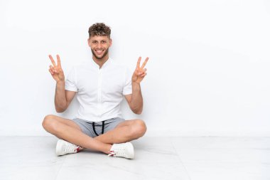 Genç sarışın adam, beyaz arka planda izole bir şekilde yerde oturuyor. İki eliyle zafer işareti gösteriyor.