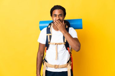 Sarı arka planda izole edilmiş büyük bir sırt çantasıyla genç dağcı adam mutlu ve ağzı eliyle kaplayan gülümsüyor.