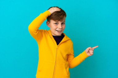 Mavi arka planda izole edilmiş küçük kızıl saçlı çocuk şaşırdı ve parmağını yana çevirdi.