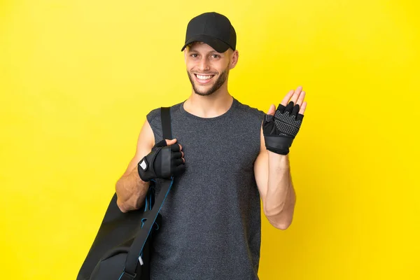 Young sport blonde man with sport bag isolated on yellow background saluting with hand with happy expression