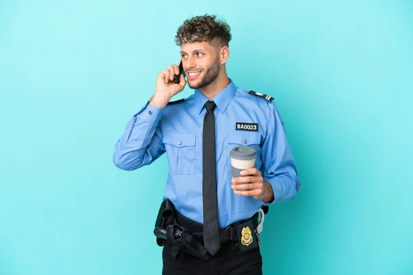 Young police blonde man isolated white on blue background holding coffee to take away and a mobile