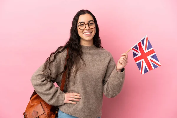 Young caucasian woman holding an United Kingdom flag isolated on pink background posing with arms at hip and smiling