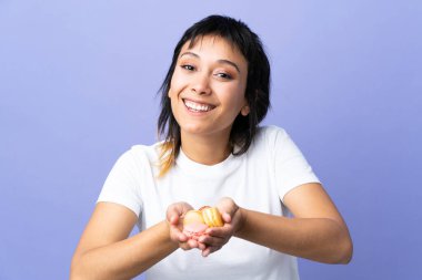 Genç Uruguaylı kadın soyutlanmış mor arka planda renkli Fransız badem kurabiyeleri tutuyor.