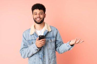 Pembe arka planda izole edilmiş cep telefonu kullanan genç Faslı adam davet edilmek için ellerini yana uzatıyor.