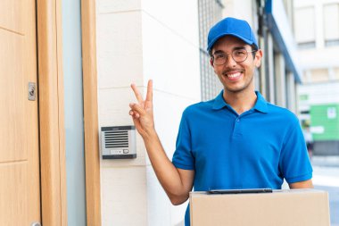 Dışarıda kutuları ve tableti tutan ve zafer jesti yapan genç teslimatçı.