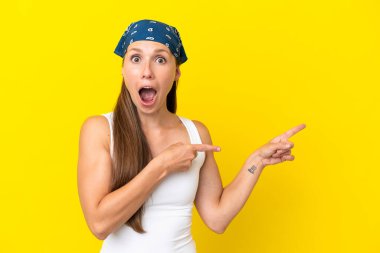 Sarı arka planda izole edilmiş genç bir İngiliz kadın şaşırmış ve işaret ediyor.