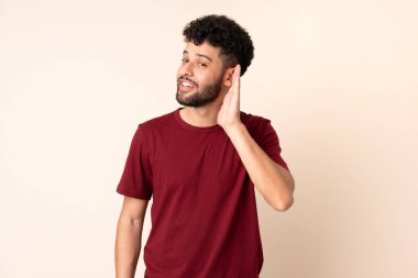 Faslı genç bir adam bej arka planda izole edilmiş. Kulağına elini sürerek bir şeyler dinliyor.