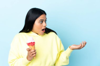 Kornet dondurması olan genç Kolombiyalı kadın mavi arka planda izole edilmiş ve yüzünde sürpriz bir ifade var.