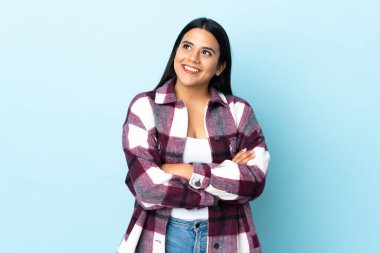 Mavi arka planda izole edilmiş genç Latin kadın gülümserken yukarı bakıyor.