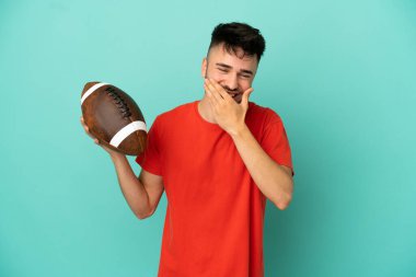 Rugby oynayan genç beyaz adam mavi arka planda izole edilmiş mutlu ve gülümseyen ağızlarını eliyle kapatıyor.