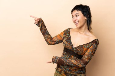 Hintli kadın bej arka planda izole edilmiş, parmağını yana doğru gösteren ve bir ürün sunan.