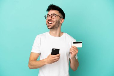 Kafkasyalı genç bir adam mavi arka planda izole edilmiş ve kredi kartıyla cep telefonu satın alıyor.