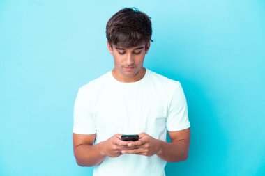 Genç beyaz adam mavi arka planda cep telefonu kullanarak izole edilmiş.