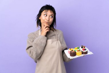 Genç Uruguaylı kadın, izole edilmiş mor arka plan üzerinde bir sürü farklı mini kek tutuyor ve bir fikir düşünüyor.