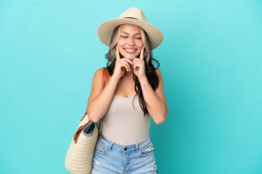 Pamel ve plaj çantalı genç Rus kız mavi arka planda izole edilmiş mutlu ve hoş bir ifadeyle gülümsüyor.