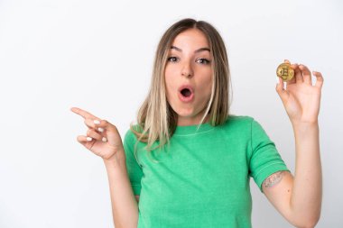 Beyaz arka planda izole edilmiş bir Bitcoin tutan genç Romen kadın şaşırmış ve işaret ediyor