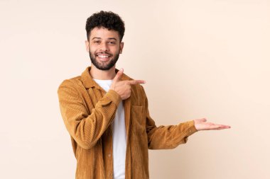 Faslı genç bir adam bej arka planda izole edilmiş. Elinde bir ilan vermek için avuç içi hayali bir şey var.