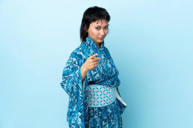 İzole edilmiş mavi arka planda kimono giyen genç bir kadın hayal kırıklığına uğramış ve ön tarafı işaret ediyor.